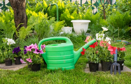 A garden scene with a neatly maintained grassy lawn foreground, featuring various potted flowers in vibrant purple and pink hues arranged along a flower bed edge. A large, green plastic watering can is positioned centrally among the flower pots, with a red-tipped spout. To the right, a small black gardening fork rests on the grass near a pink flower. In the background, lush green foliage, ferns, and mature trees create a natural backdrop, with a white garden ornament partially visible among the plants. The outdoor setting appears to be in mild weather, with bright natural light illuminating the vibrant colours of the flowers and greenery, indicating a well-tended garden space suitable for outdoor gardening and maintenance services offered by Gardening Services Charlton, serving the Charlton area within the postcode vicinity.