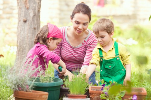 A woman and two children are participating in gardening activities in an outdoor garden space during daylight hours. The woman, dressed in a pink and white striped top, is supervising as the children, a girl in a pink headscarf and a boy in yellow and green overalls, are actively engaged in planting or tending to potted plants. The garden features a variety of flower pots and containers, some filled with leafy green plants and colorful flowers, arranged on a wooden or stone surface. In the background, there are mature trees with textured bark and green foliage, along with a blurred garden wall or fence, indicating a residential backyard or landscaped outdoor area. The scene is lit with natural sunlight, suggesting clear weather with soft shadows, contributing to a calm and inviting environment. This image exemplifies outdoor gardening activities, fostering family engagement, and maintaining a well-presented garden space, relevant for services such as gardening and lawn care offered by companies like Gardening Services Charlton in the local Charlton area.