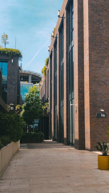 The image depicts a modern urban outdoor area adjacent to a contemporary building with a brick facade and large glass windows. A paved walkway runs alongside the building, bordered by neatly maintaine